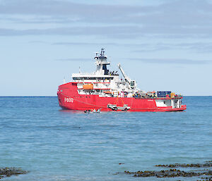 A red and white ship sits on the water jsut off shore.  A larc boat is being lowered into the water of the side of the ship.