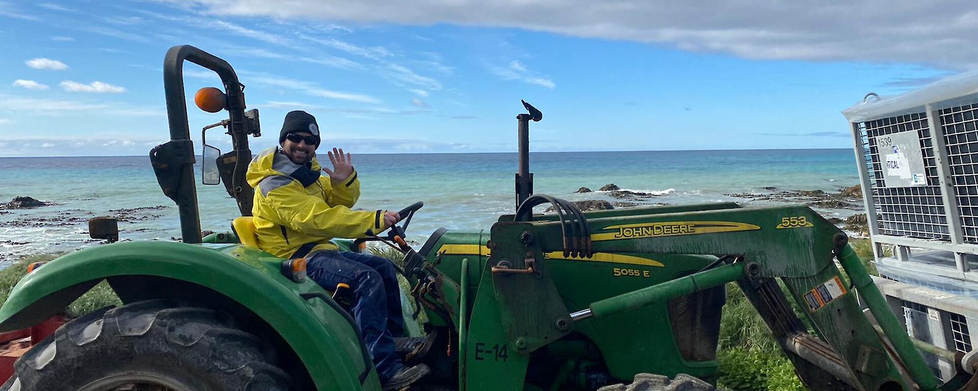 An expeditioner driving a green tractor during resupply activities, waves to camera.  The sea can be seen behind him.