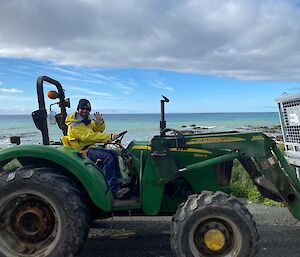 An expeditioner driving a green tractor during resupply activities, waves to camera.  The sea can be seen behind him.