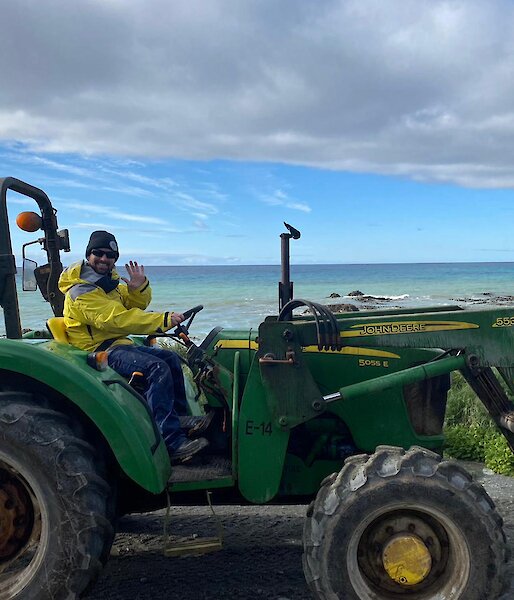 An expeditioner driving a green tractor during resupply activities, waves to camera.  The sea can be seen behind him.