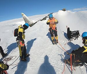 A team of expeditioners standing in the snow/ice roped together.  The tail of a crasehd aircraft, part buried in the snow can be seen behind them.