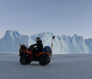 A man on a quad bike driving over the sea ice with an iceberg behind him.