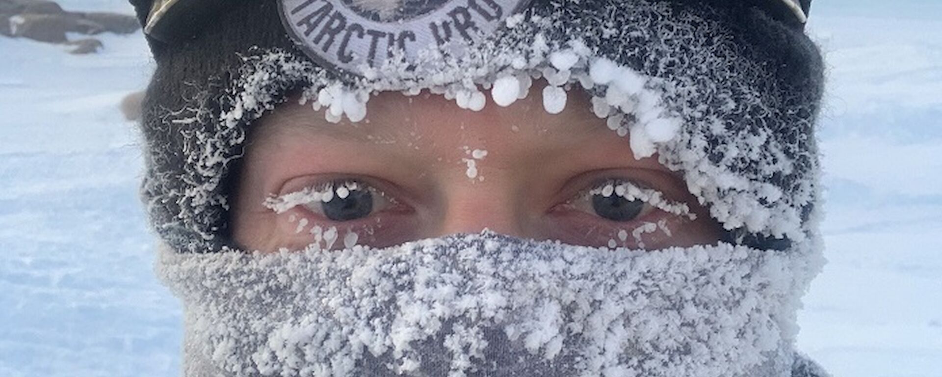 An portrait image of a man wrapped up in a beanie, snow goggles on his head and neck buff over the bottom half of his face.      Icicles are on the clothing and the ends of his eyelashes.