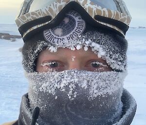 An portrait image of a man wrapped up in a beanie, snow goggles on his head and neck buff over the bottom half of his face.      Icicles are on the clothing and the ends of his eyelashes.