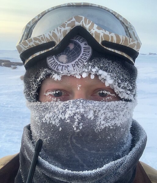 An portrait image of a man wrapped up in a beanie, snow goggles on his head and neck buff over the bottom half of his face.      Icicles are on the clothing and the ends of his eyelashes.