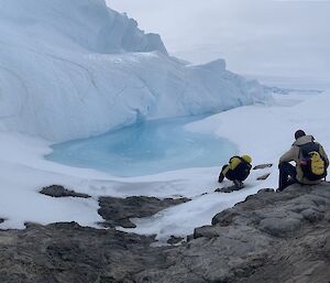 Two expeditioners with backpacks sit on the rocks near a melting glacier