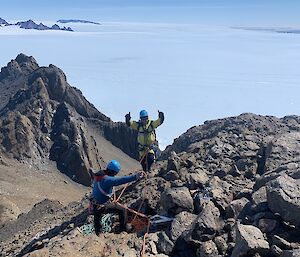 Two expeditioners stand with arms aloft at the top of a mountain.  Sea ice can be seen all around to the horizon.