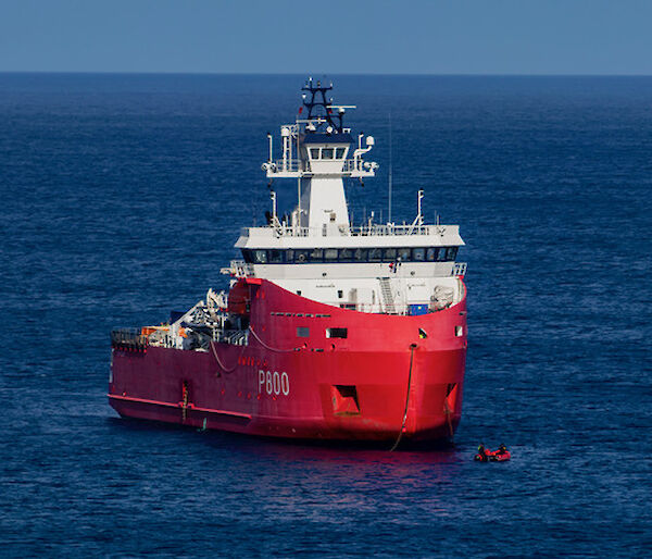 A red and white supply ship sitting in the sea with a small IRB vessel visible just of the hull