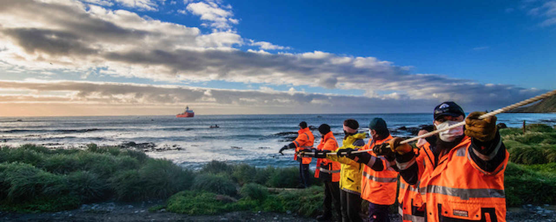 A line of expeditioners in hi-vis and wearing masks pull a fuel line in from the water to the shore.  A supply ship is visible just off shore.