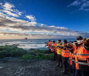 A line of expeditioners in hi-vis and wearing masks pull a fuel line in from the water to the shore.  A supply ship is visible just off shore.