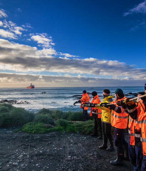 A line of expeditioners in hi-vis and wearing masks pull a fuel line in from the water to the shore.  A supply ship is visible just off shore.