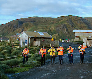 A line of expeditioners in hi-vis and wearing masks pull a fuel line in from the water to the shore.  Buildings and sheds can be seen behind them.