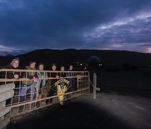 A group of expeditioners stand behind a large metal gate at dusk smiling to camera.  A sign on the gate says Welcome to Macquarie Island.