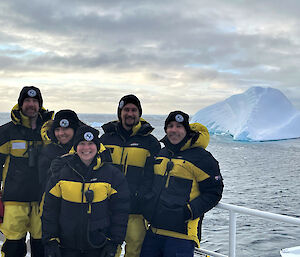 Five of the predator observer team in their yellow and black down jackets on a ship's bow with an iceberg behind.
