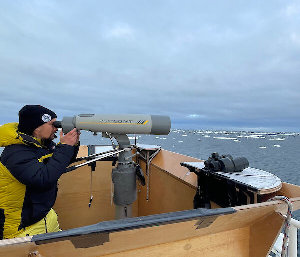 Person looking through large binoculars on bow of ship.