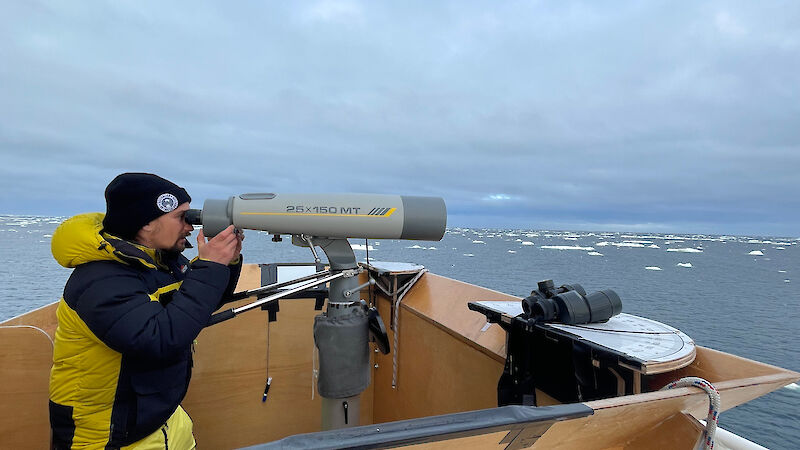 Person looking through large binoculars on bow of ship.