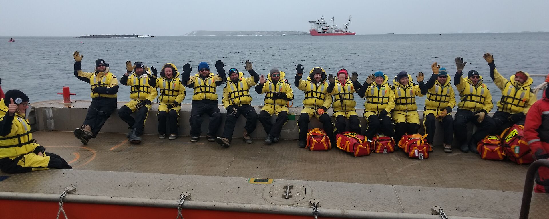 A group of expeditioners sit on the edge of a barge, waving to camera with the resupply ship in the background.