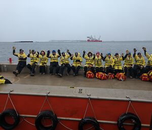 A group of expeditioners sit on the edge of a barge, waving to camera with the resupply ship in the background.