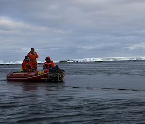 3 expeditioners in a dinghy sit on the water watching a fuel line.  Icebergs in the distance.