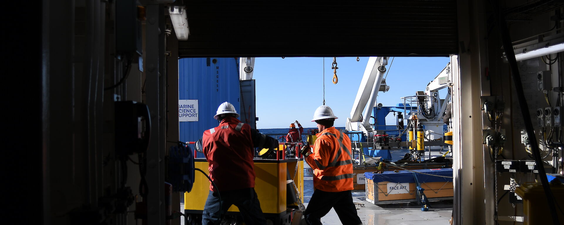 Two people push a large, square mooring across the deck of a ship.