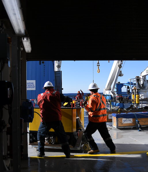 Two people push a large, square mooring across the deck of a ship.