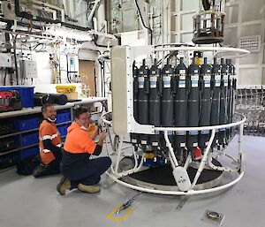 Two men on a ship with a large, circular water sampling instrument, containing a lot of bottles.