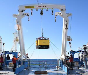 A large, square, yellow, box-shaped mooring hoisted over the back of a ship, ready to be deployed into the ocean.
