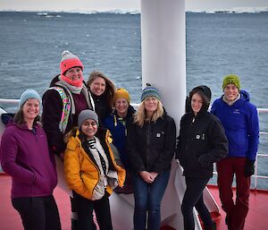 A group of women in beanies and warm jackets standing smiling to camera at the back of a ship