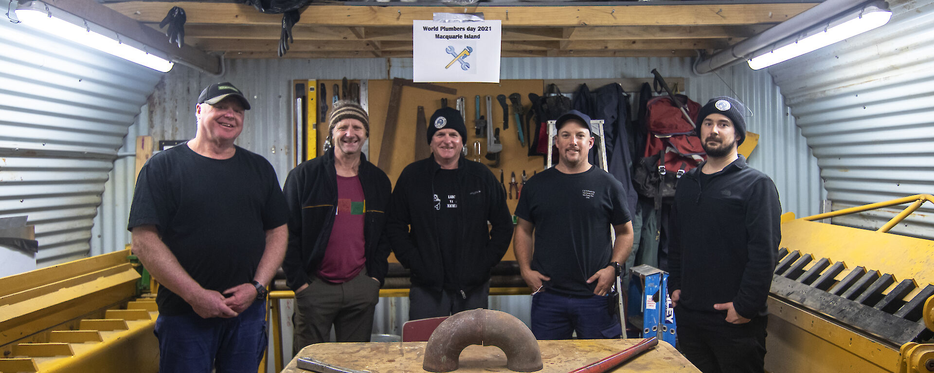 5 men stand behind a work bench facing camera and smiling