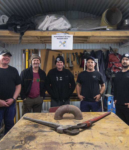 5 men stand behind a work bench facing camera and smiling