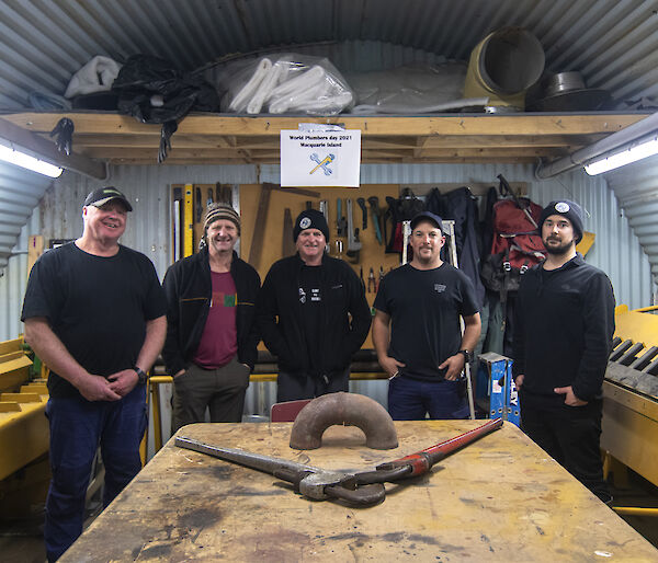 5 men stand behind a work bench facing camera and smiling