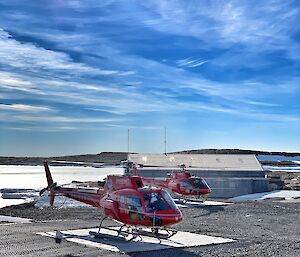 Two helicopters on the ground with a shed and icy ground in the background