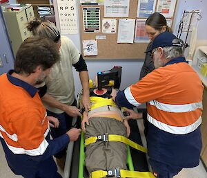 Four people, two in hi-vis, stand around a dummy lying on a gurney.  Two of the people are strapping the dumming in.