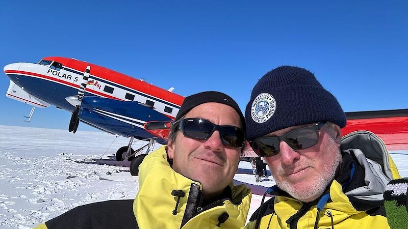 Two men in yellow jackets standing in front of a red, white and blue Basler aircraft in Antarctica.