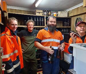 Four men, three in hi-vis standing in a shed with shelves of brown ale bottles on the shelves.  A couple of the men are holding some bottles.  All are smiling to camera.