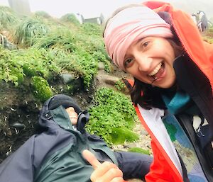 A woman close to camera smiling with a thumbs up, pointing at a dummy, dressed in hiking gear, lying on the ground