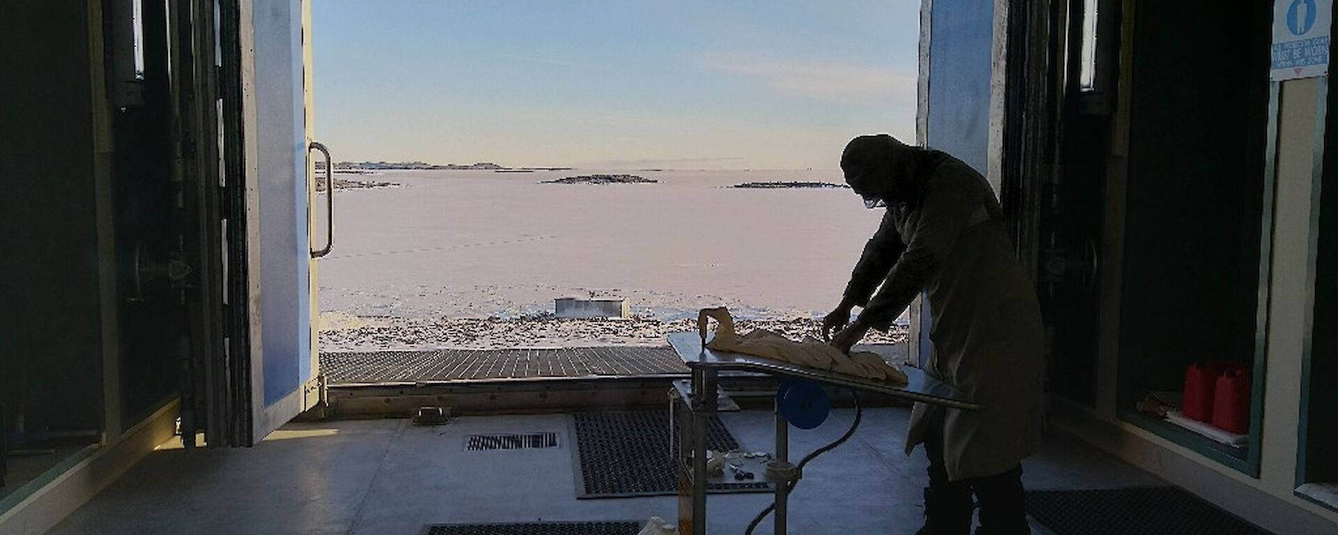A man stands in a hut prepares a weather balloon for inflation at a small table.   Large doors are open and the Antarctic landscape can be seen behind the man.