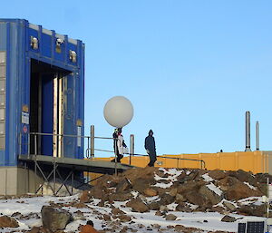 Two people come out of a blue shed and are walking down a ramp.  One holds a giant inflated weather balloon