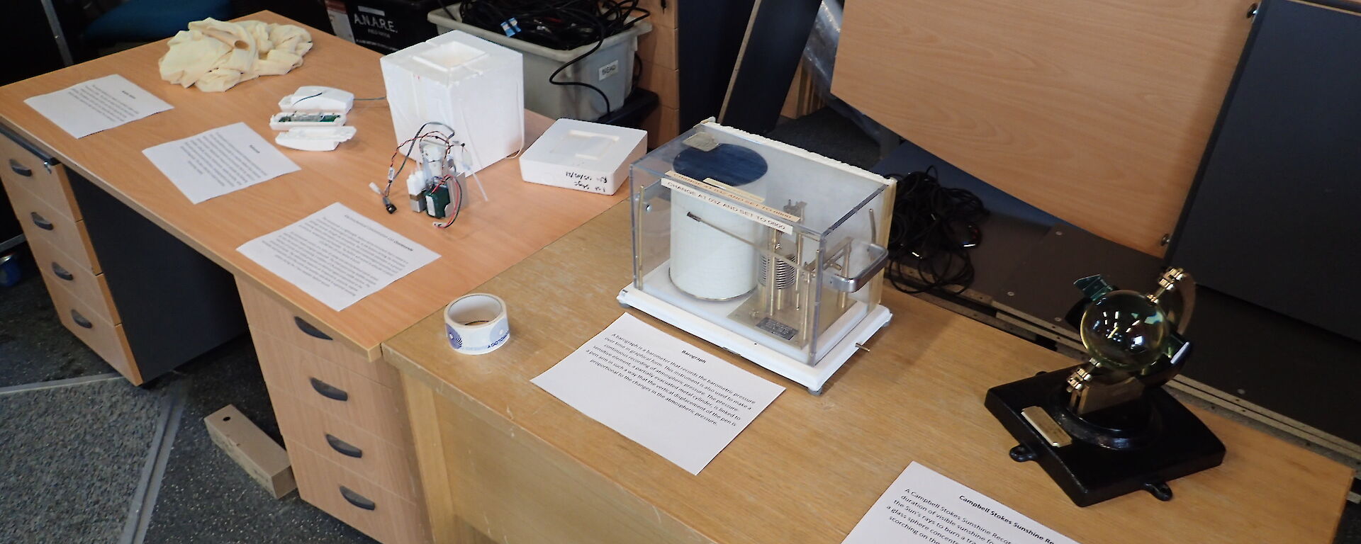 Two desks pushed together with meteorological instruments displayed with sheets of paper in front which explain what the equipment is for.