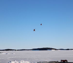 Two red helicopters flying low over the sea ice as they approach the landing pad.