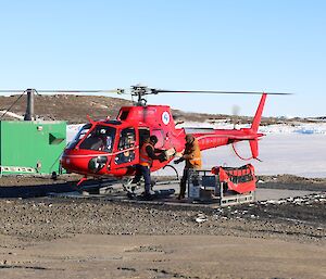 Red helicopter on the landing pad while two people unload the cargo into a cage pallet.