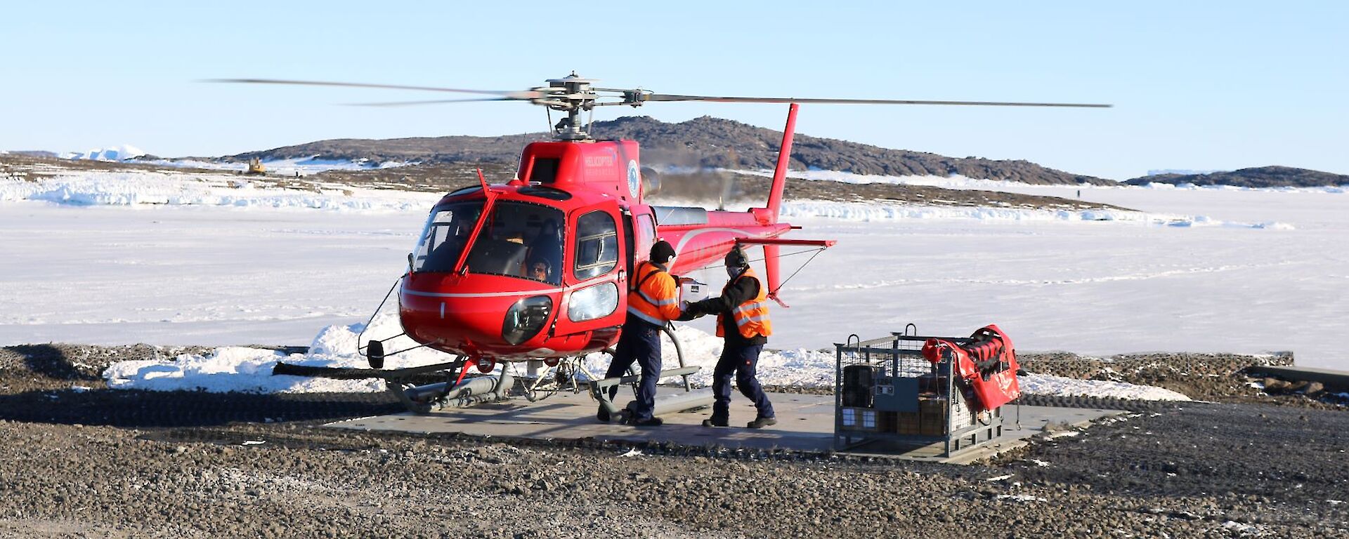 Red helicopter on the landing pad while two people unload the cargo into a cage pallet.