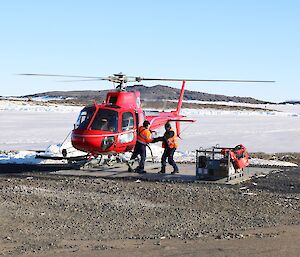 Red helicopter on the landing pad while two people unload the cargo into a cage pallet.