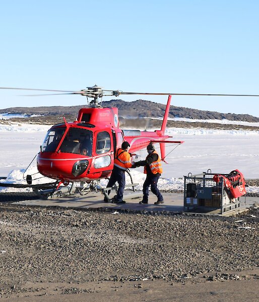 Red helicopter on the landing pad while two people unload the cargo into a cage pallet.