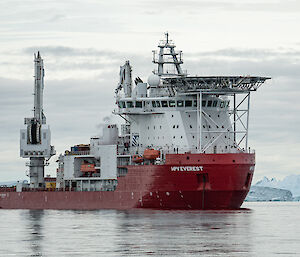 A large red ship sits in the calm water with large icebergs visible behind