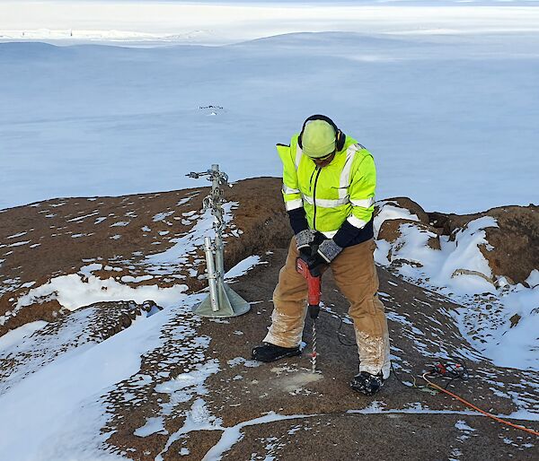 Casey expeditioner drilling a hole in the snow covered rocks for weather instruments