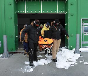 People caring an orange Ferno stretcher out of a large green building.