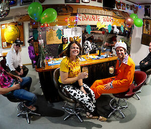 Some people sitting at a bar smiling to camera in fancy dress.  The bar is decorated with balloons.