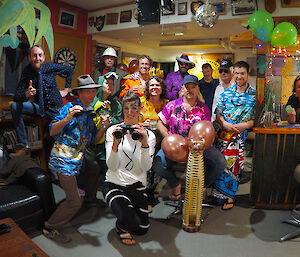 A group shot of expeditioners all dressed up as characters from the film Madagascar.  The room is decorated with palm trees and balloons.