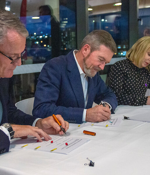 Three people signing paperwork, seated at a table.